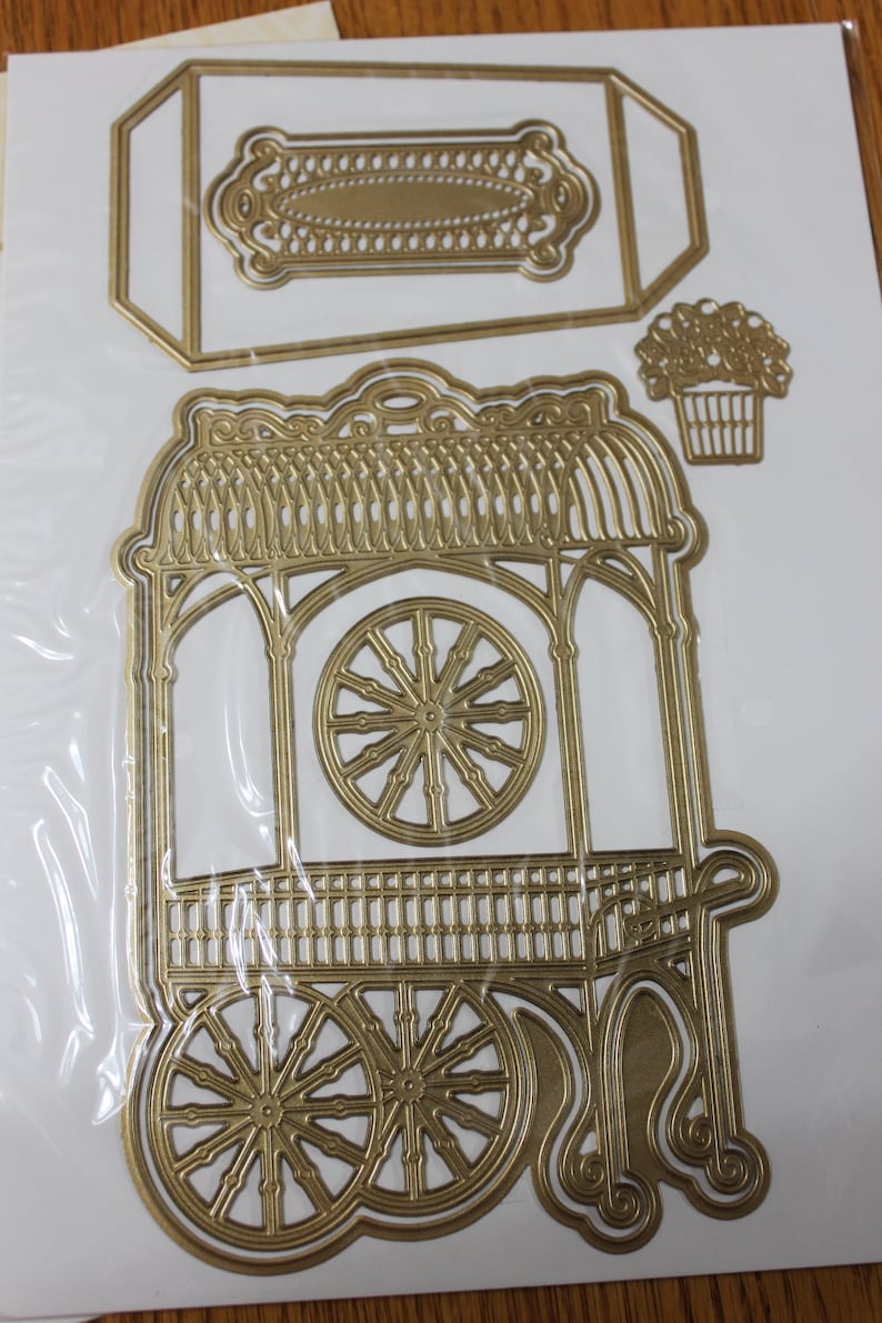 May include: A gold metal die-cut set of a decorative cart with a wheel, a rectangular sign, and a flower pot. The cart has a decorative roof and a lattice-style side.