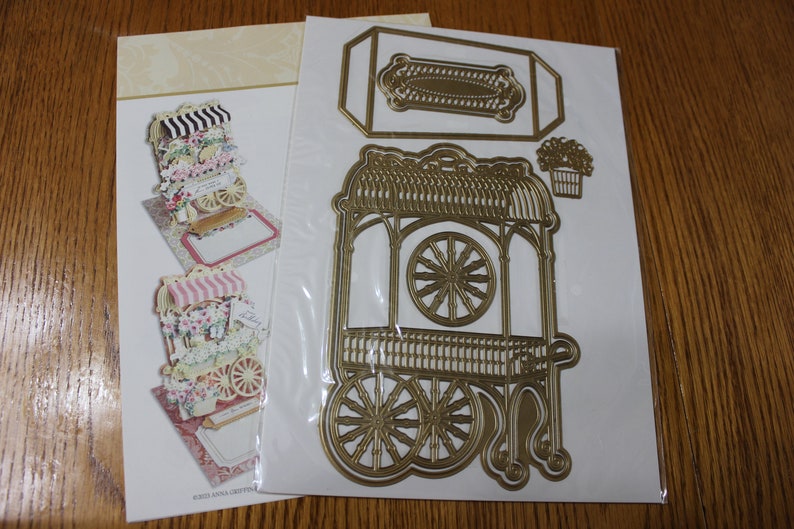May include: A gold metal die-cut set for crafting, featuring a flower cart with a canopy, wheels, and a planter. The set includes a separate die-cut for a rectangular sign.