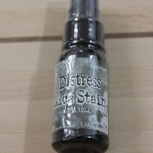 May include: A small, clear bottle with a black spray top. The bottle is labeled "Distress Mica Stain - Dusty Tomb - 1 fl oz (29 ml)"