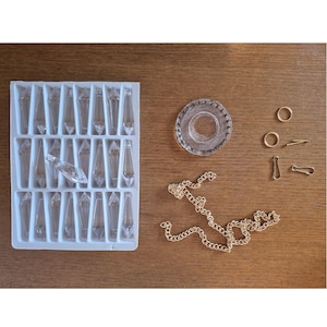 May include: A white plastic tray containing twenty-one clear, faceted, teardrop-shaped glass crystals. Beside the tray are a circular glass component, a gold-toned metal chain, two small gold-toned rings, and two gold-toned metal lobster clasps.