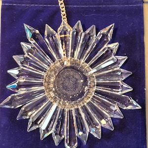 May include: A clear, faceted crystal suncatcher shaped like a star or flower with pointed rays radiating from a circular center. The crystal ornament is suspended by a thin gold-toned metal chain and rests against a dark blue velvet pouch.