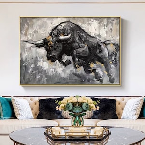 May include: A black and white abstract painting of a bull charging forward with gold accents. The painting is framed in a gold frame and is hanging on a white wall above a couch.