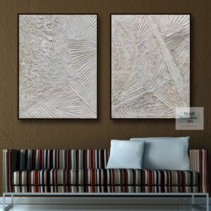May include: Two white abstract paintings with textured lines and shapes. The paintings are framed in black and are hanging on a brown wall above a striped couch.