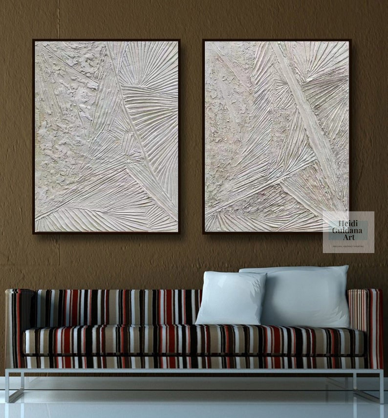 May include: Two white abstract paintings with textured lines and shapes. The paintings are framed in black and are hanging on a brown wall above a striped couch.