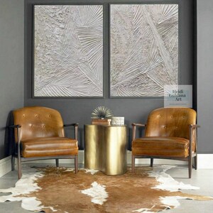 May include: Two abstract white textured paintings with a gray background, hanging above two brown leather chairs and a gold metal side table. The text "Heidi Guidana Art" is visible in the bottom right corner of the image.
