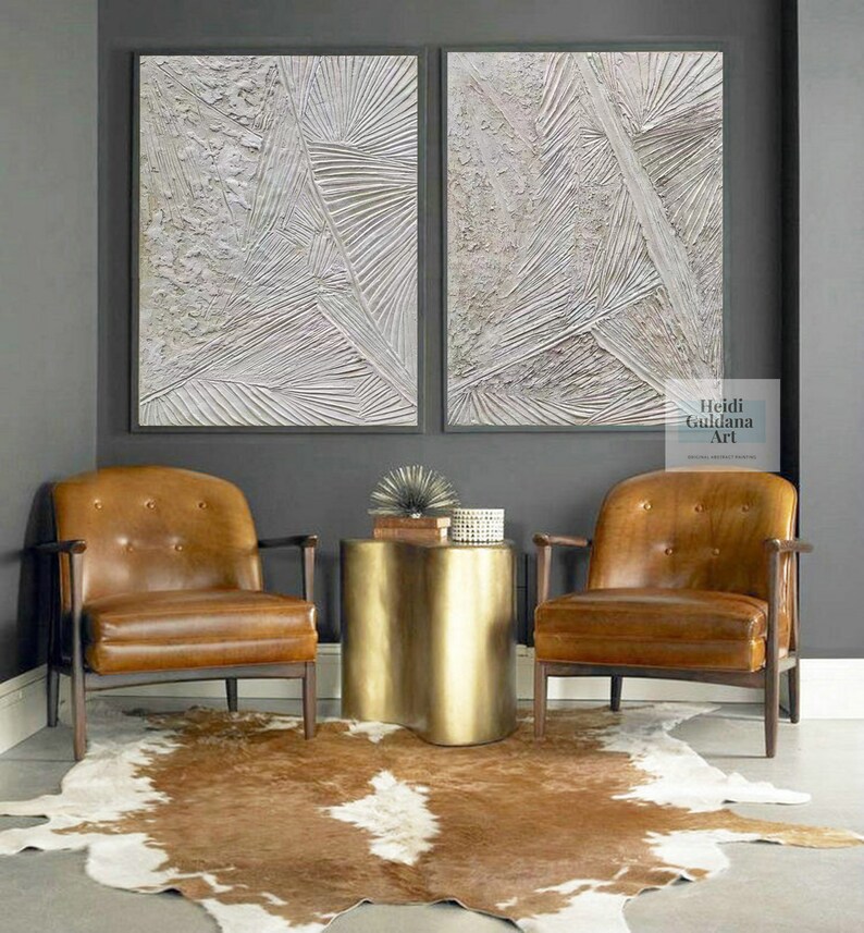 May include: Two abstract white textured paintings with a gray background, hanging above two brown leather chairs and a gold metal side table. The text "Heidi Guidana Art" is visible in the bottom right corner of the image.