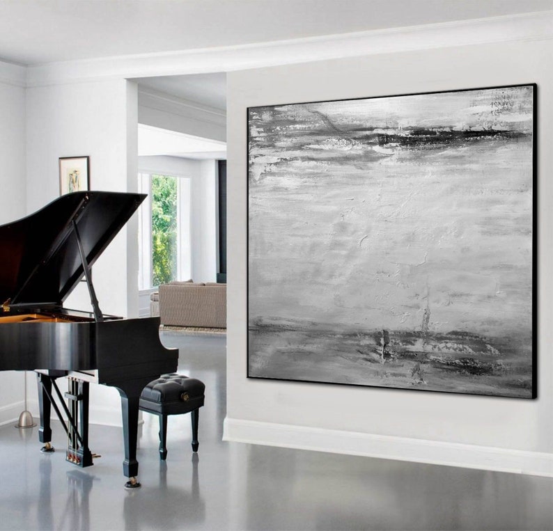 Op de afbeelding: Een abstract zwart-wit schilderij met een horizontale compositie. Het schilderij heeft een textuur, gelaagd effect met tinten grijs en wit. Het schilderij is ingelijst in een zwart frame en hangt aan een witte muur.