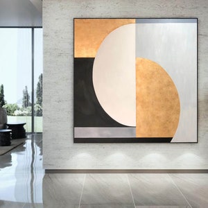 May include: Abstract geometric painting with a large white circle, black and gold sections, and a silver stripe. The painting is framed and hanging on a gray wall.