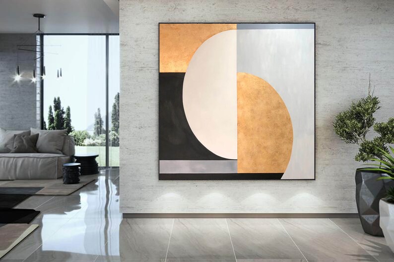 May include: Abstract geometric painting with a large white circle, black and gold sections, and a silver stripe. The painting is framed and hanging on a gray wall.