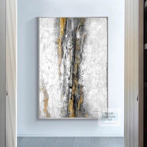 May include: Abstract painting with gold, black, and white paint on a white canvas. The painting has a textured look and is framed in a thin, white frame.