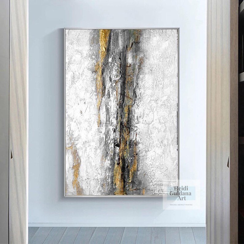 May include: Abstract painting with gold, black, and white paint on a white canvas. The painting has a textured look and is framed in a thin, white frame.