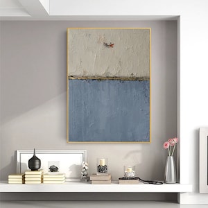 May include: Abstract painting with a textured white background and a blue bottom half. The painting is framed in gold and has a thin gold line separating the two colors.