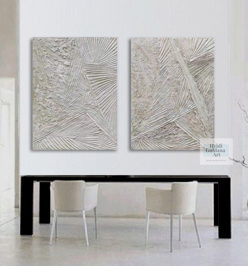 May include: Two abstract paintings in white and gray tones with textured lines and shapes. The paintings are hung on a white wall above a black table with two white chairs.