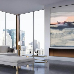 May include: A modern living room with a large abstract painting in shades of gray, blue, and pink hanging above a fireplace. The painting is framed in black and the fireplace is made of white stone. The room has large windows with a view of a city skyline. There is a white sofa and two white ottomans in the room.