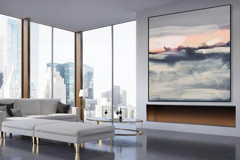 May include: A modern living room with a large abstract painting in shades of gray, blue, and pink hanging above a fireplace. The painting is framed in black and the fireplace is made of white stone. The room has large windows with a view of a city skyline. There is a white sofa and two white ottomans in the room.
