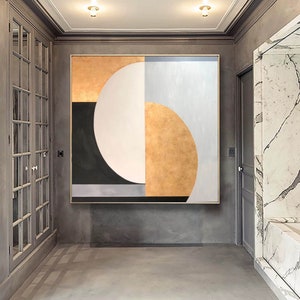 May include: Abstract geometric painting with a large white circle, a black crescent shape, and gold and gray sections. The painting is framed and hanging on a gray wall.