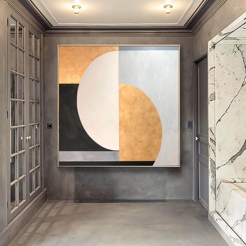 May include: Abstract geometric painting with a large white circle, a black crescent shape, and gold and gray sections. The painting is framed and hanging on a gray wall.