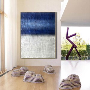 Puede incluir: Pintura abstracta con un esquema de color azul y blanco. La pintura est&aacute; colgada en una pared en una casa moderna con paredes blancas y un suelo de madera. Hay tres pufs tejidos en primer plano.