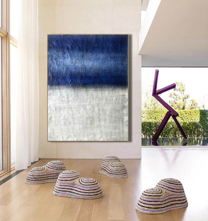 Puede incluir: Pintura abstracta con un esquema de color azul y blanco. La pintura est&aacute; colgada en una pared en una casa moderna con paredes blancas y un suelo de madera. Hay tres pufs tejidos en primer plano.