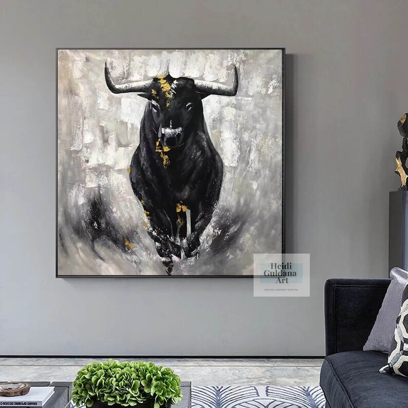 May include: A large square canvas painting depicts a black bull charging forward. The bull has large horns and gold accents on its head and body. The background is a textured mix of gray and white brushstrokes, framed in a black frame.