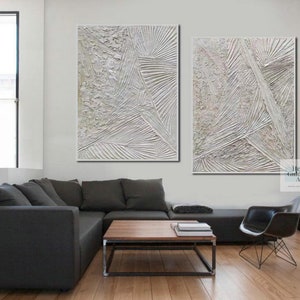 May include: Two abstract paintings in white and gray tones with textured lines and patterns. The paintings are hung on a white wall above a gray sectional sofa and a coffee table.