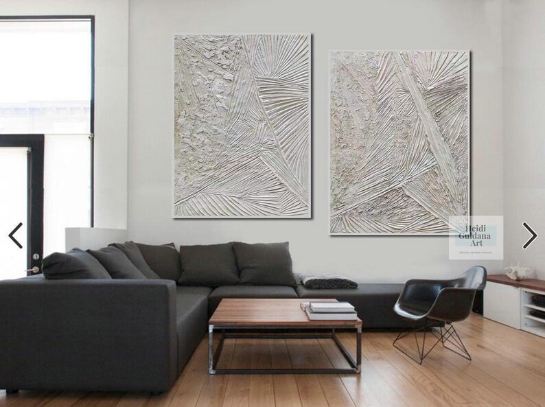 May include: Two abstract paintings in white and gray tones with textured lines and patterns. The paintings are hung on a white wall above a gray sectional sofa and a coffee table.