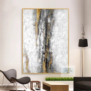 May include: Abstract painting with gold and gray accents in a gold frame. The painting is on a white wall with a black lamp and a chair in the background. The artist's name, Heidi Guldana Art, is in the bottom right corner of the painting.
