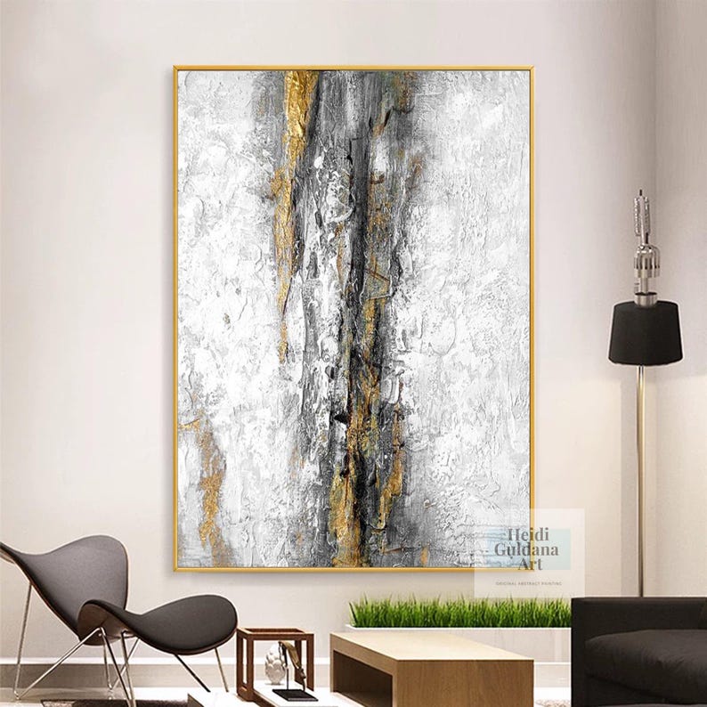 May include: Abstract painting with gold and gray accents in a gold frame. The painting is on a white wall with a black lamp and a chair in the background. The artist's name, Heidi Guldana Art, is in the bottom right corner of the painting.