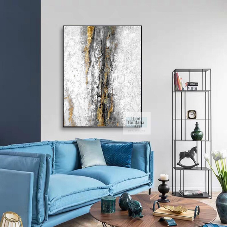 May include: Abstract painting with a white background and gold and gray accents. The painting is hanging on a white wall above a blue sofa.