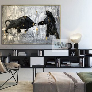 May include: Abstract painting of a black bull and bear facing each other on a gray and gold background. The painting is framed in gold and hanging on a wall in a modern living room.