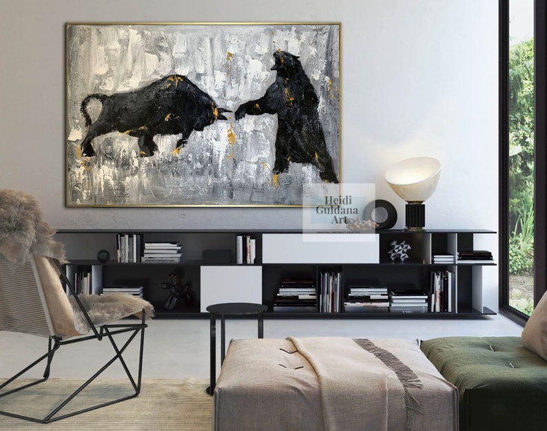 May include: Abstract painting of a black bull and bear facing each other on a gray and gold background. The painting is framed in gold and hanging on a wall in a modern living room.