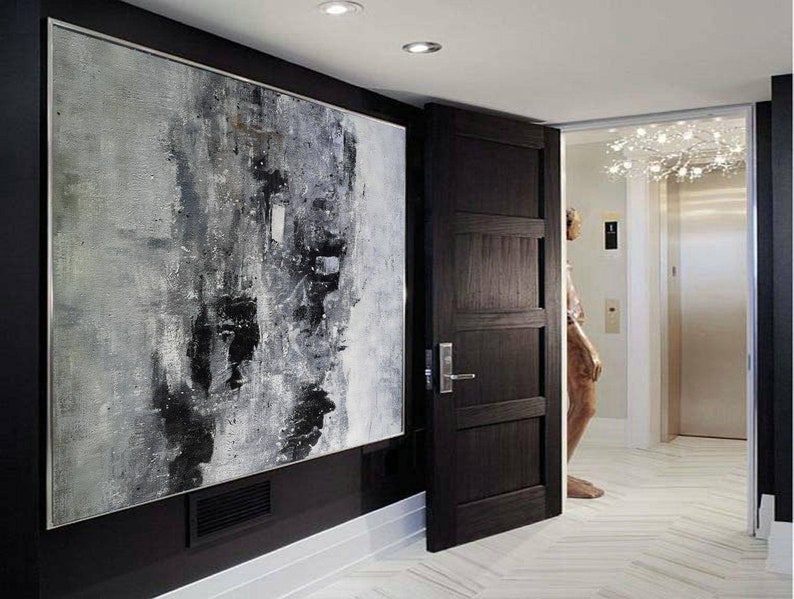 May include: A black and white abstract painting hangs on a black wall in a modern hallway. A dark wood door is open to reveal a white tiled floor and a hallway with a chandelier.