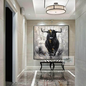 May include: Large black and white bull painting with gold accents. The artwork is displayed in a hallway with a black frame. The painting is above a black and white striped bench. The artist's name is in the bottom right corner.