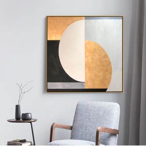 May include: Abstract geometric painting with a gold, white, black, and gray color scheme. The painting features a large white circle with a gold crescent moon shape on the right side. The painting is framed in a gold frame.
