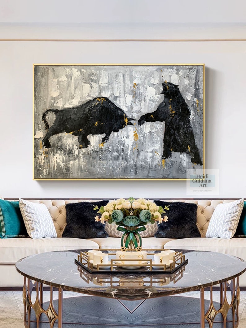 May include: A black and white abstract painting of a bull and a bear facing each other. The painting is on a canvas with a white background and gold accents. The painting is hanging on a wall above a couch with pillows and a coffee table.