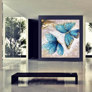 May include: A painting of two blue butterflies with gold accents on a white background. The butterflies are in flight and their wings are spread wide. The painting is framed in a black frame and is hanging on a wall.