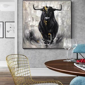 May include: A large, framed canvas art piece featuring a black bull with gold accents against a textured gray and white background. The bull is the focal point, with its horns and muscular form prominently displayed. The artwork is a modern, abstract representation.