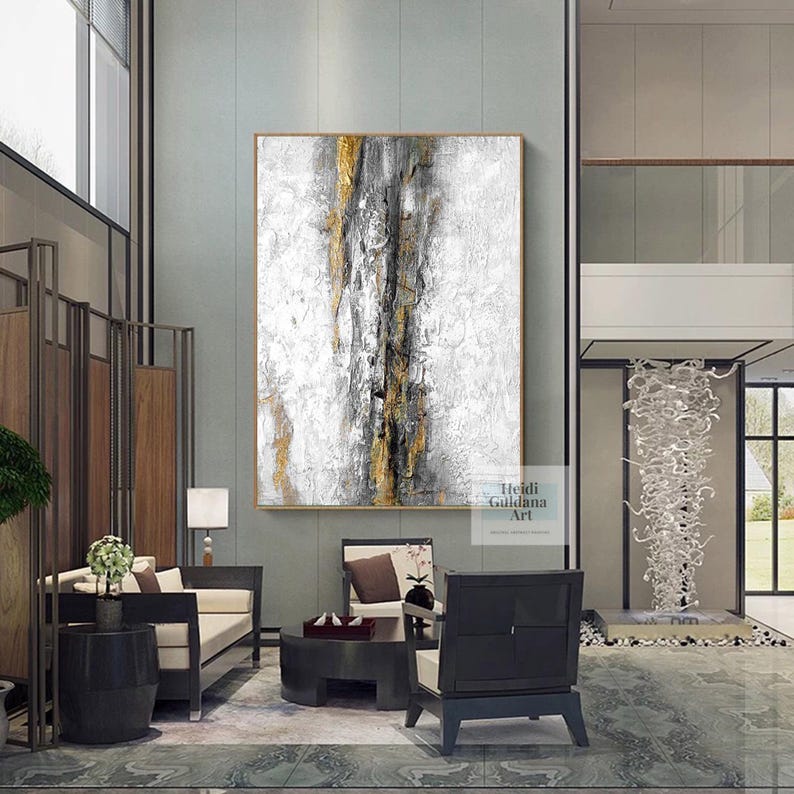 May include: Abstract painting with gold, black, and white paint on a canvas. The painting is hanging on a wall in a modern living room.