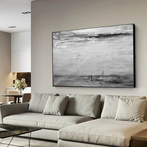 Op de afbeelding: Een zwart-wit abstract schilderij met horizontale penseelstreken. Het schilderij is ingelijst in een zwart frame en hangt aan een muur boven een witte bank.