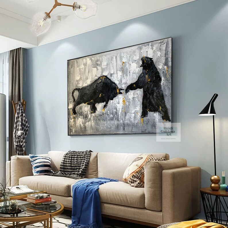 May include: Abstract painting of a black bull and bear facing each other on a white and gray background with gold accents. The painting is framed and hanging on a wall in a living room.