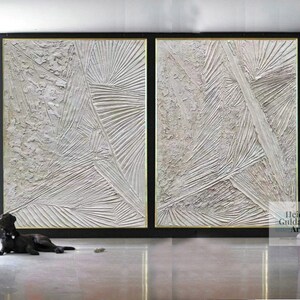 May include: Two large abstract paintings in white with textured lines and a gold frame. The paintings are hanging on a black wall in a modern interior.