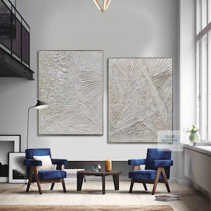 May include: Two abstract paintings in white and gray with textured lines and shapes. The paintings are framed and hung on a white wall in a modern living room.