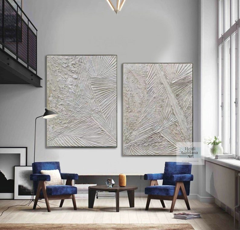 May include: Two abstract paintings in white and gray with textured lines and shapes. The paintings are framed and hung on a white wall in a modern living room.