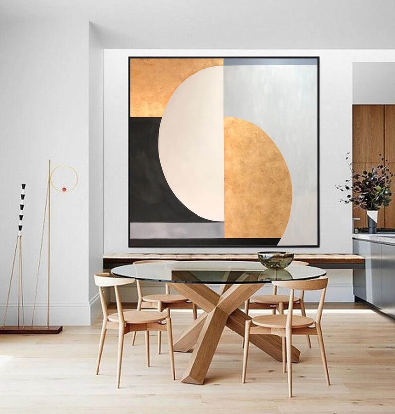 May include: Abstract geometric painting with gold, white, black, and gray colors. The painting features a large white circle with a gold crescent shape on the right side. The painting is hanging on a white wall above a wooden table with four chairs.