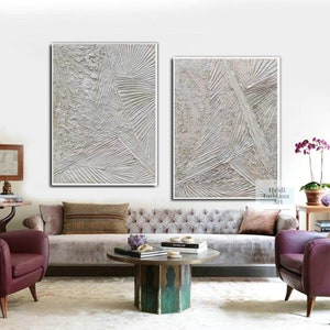 May include: Two abstract paintings in white and gray tones with textured lines and patterns. The paintings are framed in white and are hanging on a white wall above a gray sofa.
