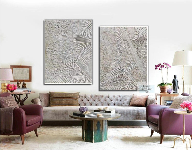 May include: Two abstract paintings in white and gray tones with textured lines and patterns. The paintings are framed in white and are hanging on a white wall above a gray sofa.
