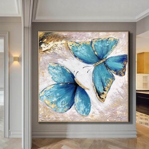 May include: A painting of two blue butterflies with gold accents on a white background. The butterflies are painted in a textured style, giving the painting a three-dimensional effect.
