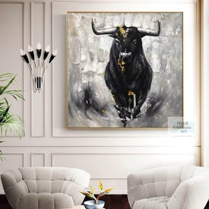 May include: A large square painting featuring a black bull charging forward. The bull has gold accents and is set against a gray and white abstract background. The artwork is framed in gold and hangs on a white wall.