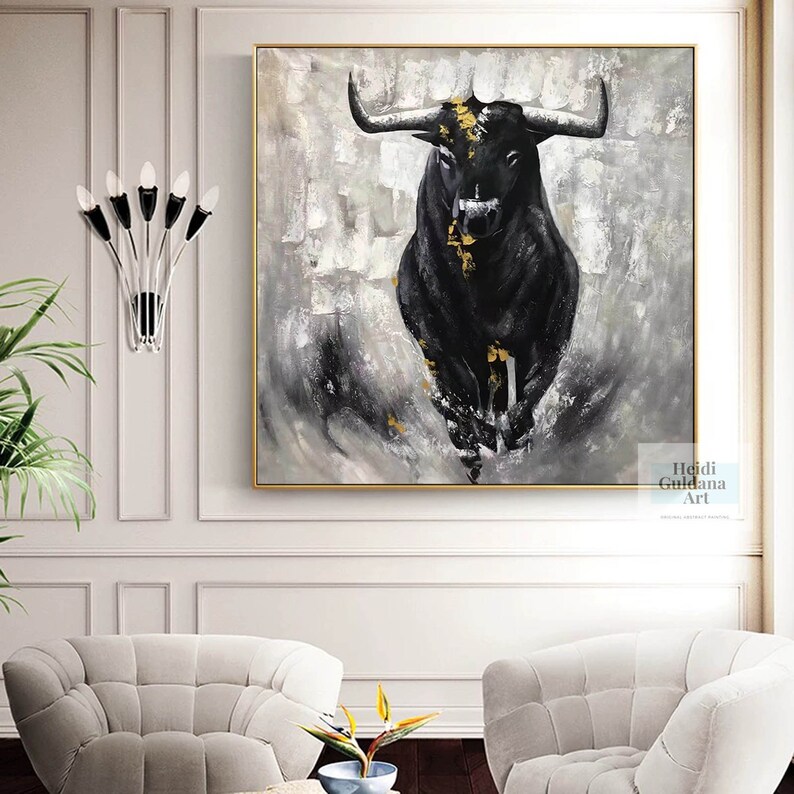 May include: A large square painting featuring a black bull charging forward. The bull has gold accents and is set against a gray and white abstract background. The artwork is framed in gold and hangs on a white wall.