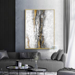 May include: Abstract painting with gold, gray, and white colors. The painting is framed in a gold frame and is hanging on a gray wall.